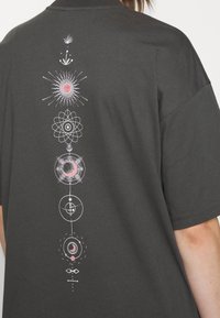 Una serie de símbolos celestiales en blanco y rosa, que incluyen el sol, la luna y diseños atómicos, impresos verticalmente en la parte posterior de una camiseta gris oscuro.