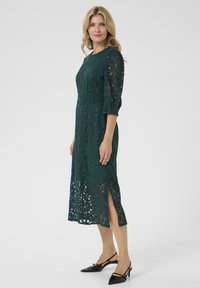 Türkiz csipke midi ruha háromnegyedes ujjal, kerek nyakkivágással és oldalsó hasítékokkal; fekete, sarkú slingback magassarkúkkal párosítva.