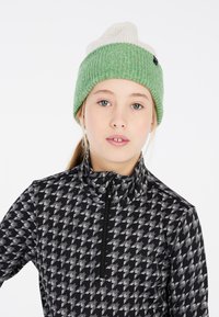 Bonnet en tricot avec un schéma de couleurs vert et crème, présentant un design en côtes texturé. Le modèle porte un haut zippé à motifs noir et gris.