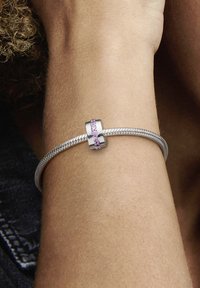 Silberarmband mit strukturiertem Band und einem zentralen Verschluss, der mit rosa Edelsteinen in einem kreisförmigen Design verziert ist. Am Handgelenk getragen.
