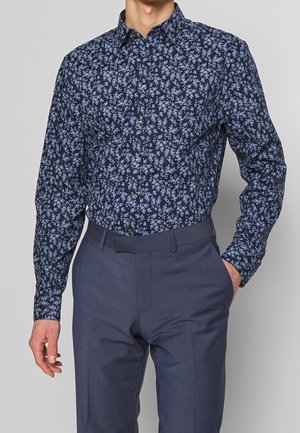 Homme portant une chemise bleu marine à manches longues avec un petit imprimé floral bleu clair et un pantalon de costume gris foncé, main dans la poche et fond neutre.