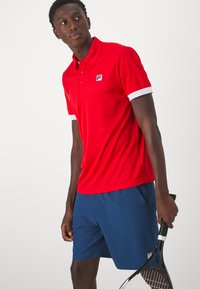 Polo rouge avec des accents de col blanc et un logo, associé à un short bleu. Le modèle tient une raquette de tennis dans une pose neutre.