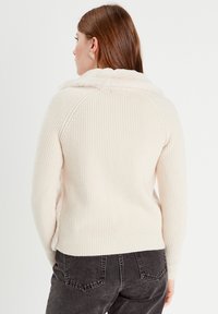 Suéter de canalé crema con un suave y esponjoso cuello, que cuenta con mangas largas y un diseño ajustado. Combinado con pantalones gris oscuro.