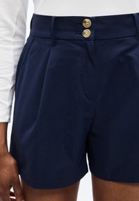 Σορτς σε σκούρο μπλε ναυτικού ύφους με ψηλή μέση, δύο μπεζ κουμπιά, πλαϊνές τσέπες και πιέτες, που φοριέται με λευκό μακρυμάνικο τοπ.