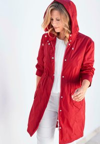 Rote leichte Jacke mit Kapuze, elastischem Bund, vorderem Reißverschluss und Druckknopfdetails. Zwei aufgesetzte Taschen vorne.