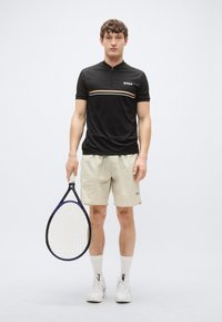 Svart kortermet polo med glidelåsåpning i kragen og beige shorts, holder en tenniskølle. Har "BOSS"-logo og fargeaksenter. Hvite sneakers.