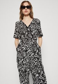 Combinaison noire et blanche à motifs avec des manches courtes, décolleté en V, taille cintrée et poches avant, présentant un design graphique et abstrait.