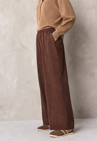 Braune Hosen mit weitem Bein und elastischem Bund, aus weichem Stoff gefertigt. Kombiniert mit einem beigefarbenen Pullover und braunen Sneakers.