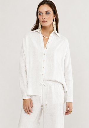 Vrouw draagt een losse witte blouse met knopen en bijpassende broek met trekkoord, lange kralenoorbellen en een schelpenketting tegen een effen achtergrond.