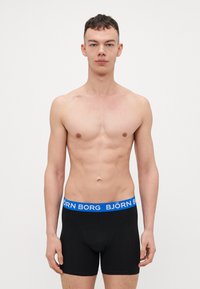 Fekete boxer alsónadrág kék derékkal, rajta a "BJÖRN BORG" felirattal. Az anyag lágy, rugalmas és testhez simuló kinézetű. A modellen izmos testalkat látható.