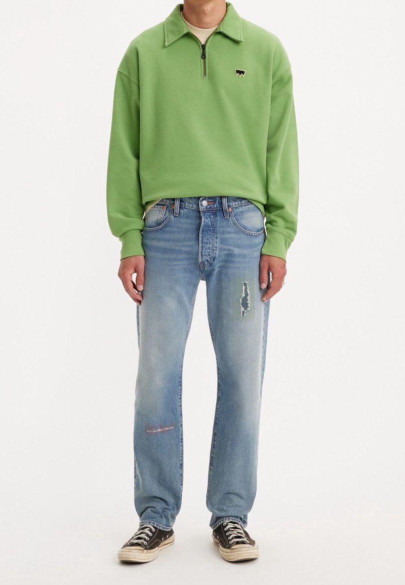 Felpa verde con mezza zip e piccolo logo, abbinata a jeans azzurri chiari strappati con una toppa tessuta e dettagli sfrangiati, indossata con sneakers nere.