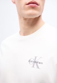 Κοντινό πλάνο ενός ατόμου που φοράει λευκό πουκάμισο με μικρό λογότυπο Calvin Klein στο στήθος, εμφανίζοντας τα χείλη, το σαγόνι και τον λαιμό.