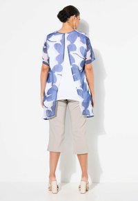 Helle, leichte blau-blumige Overlay-Bluse mit kurzen Ärmeln, offenem Rücken und weißem Untershirt, kombiniert mit beigen, kurzen Hosen.