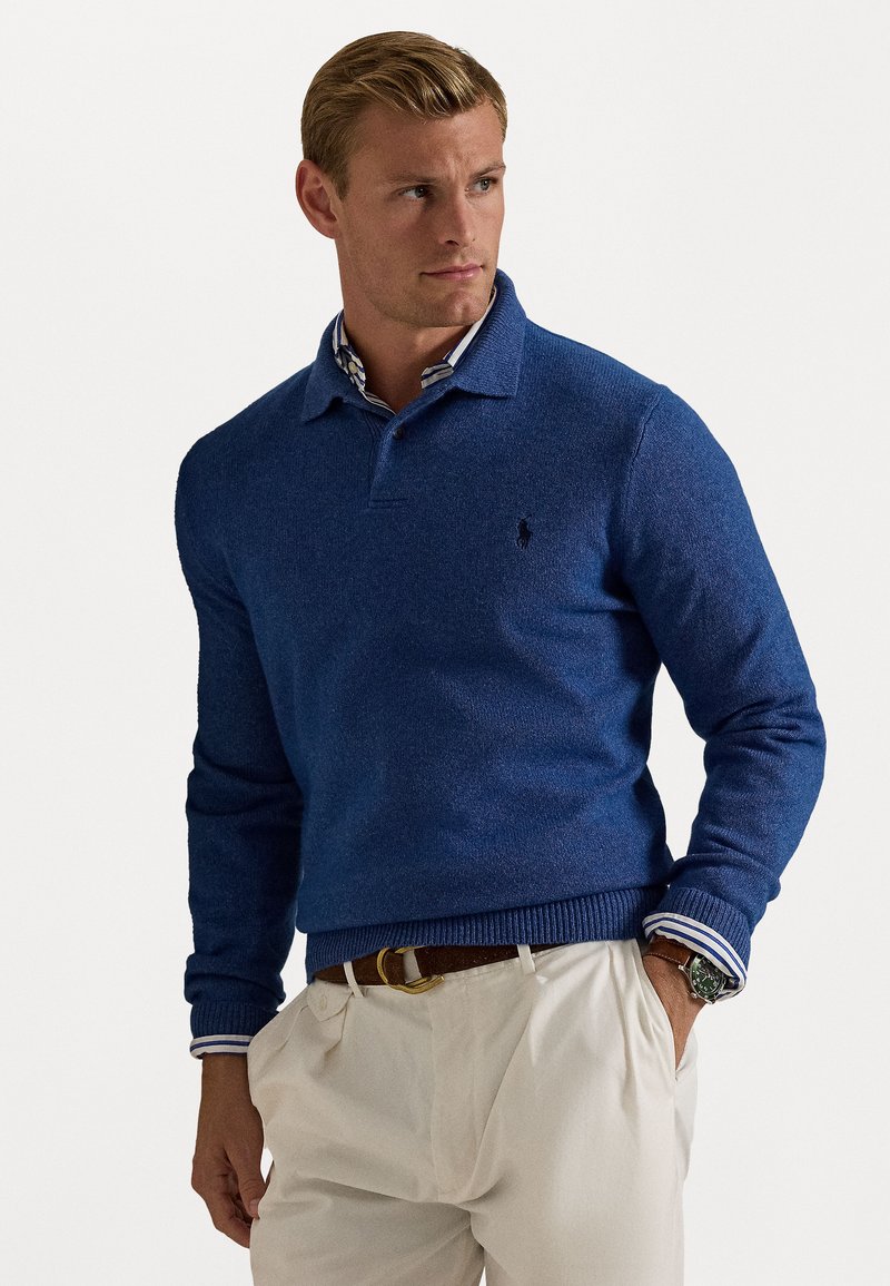 Blåt strikket sweater med polo-krave og ribbet kant. Broderet logo på brystet. Laget over en hvid skjorte med blå striber.