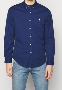 Man med en mörkblå skjorta med lång ärm och knappar, med en liten gul logotyp på bröstet, ihop med ljusblå jeans.