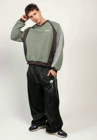 En ung man med kort hår som bär en grön och grå sweatshirt, lösa svarta byxor och sneakers, står med en hand bakom huvudet.