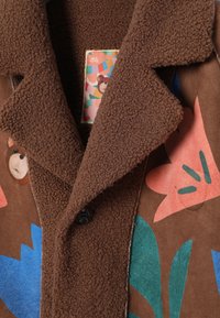 Brązowy płaszcz z barankiem z kolorowymi twarzami niedźwiedzi i abstrakcyjnymi, różowymi, niebieskimi oraz zielonymi kwiatowymi wzorami na panelach materiału.