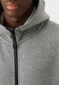 Nahaufnahme einer Person, die einen grauen Kapuzenpullover mit Reißverschluss und hohem Kragen trägt, mit dezentem „hummel“-Logo auf der Brust.