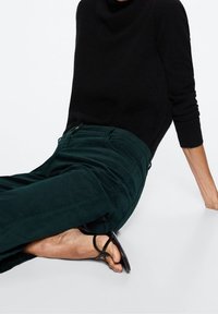 Mustita neulepaita, joka on löysästi istuva, yhdistetty tummaansateenharmaisiin samettihousuihin. Jalkoja koristaa yksinkertaiset mustat sandaalit.