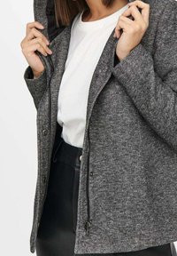 Grå strikket jakke med en struktureret finish, lynlås foran og trykknapper. Bæres over en hvid skjorte, har en hætte.