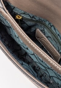 Zbliżenie na otwartą torbę ze skóry w kolorze taupe, ukazującą wnętrze z niebieskim materiałem w wzory oraz złoty suwak na kieszeni zapinanej na zamek.