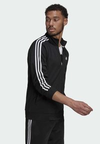 Chaqueta deportiva negra con un diseño de tres rayas blancas en las mangas, cremallera completa, cuello alto y puños elásticos. Llevada sobre una camisa blanca.