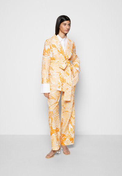 Conjunto de duas peças com estampado floral amarelo e branco, incluindo um casaco com cinto e calças de perna larga com mangas compridas e detalhes brancos. Sandálias transparentes.
