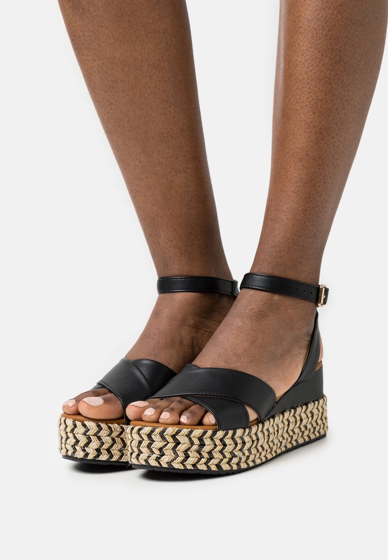 Sandales à plateforme noires avec des brides croisées, une semelle intermédiaire en jute tressé et une bride à la cheville. Surface texturée et design à bout ouvert.