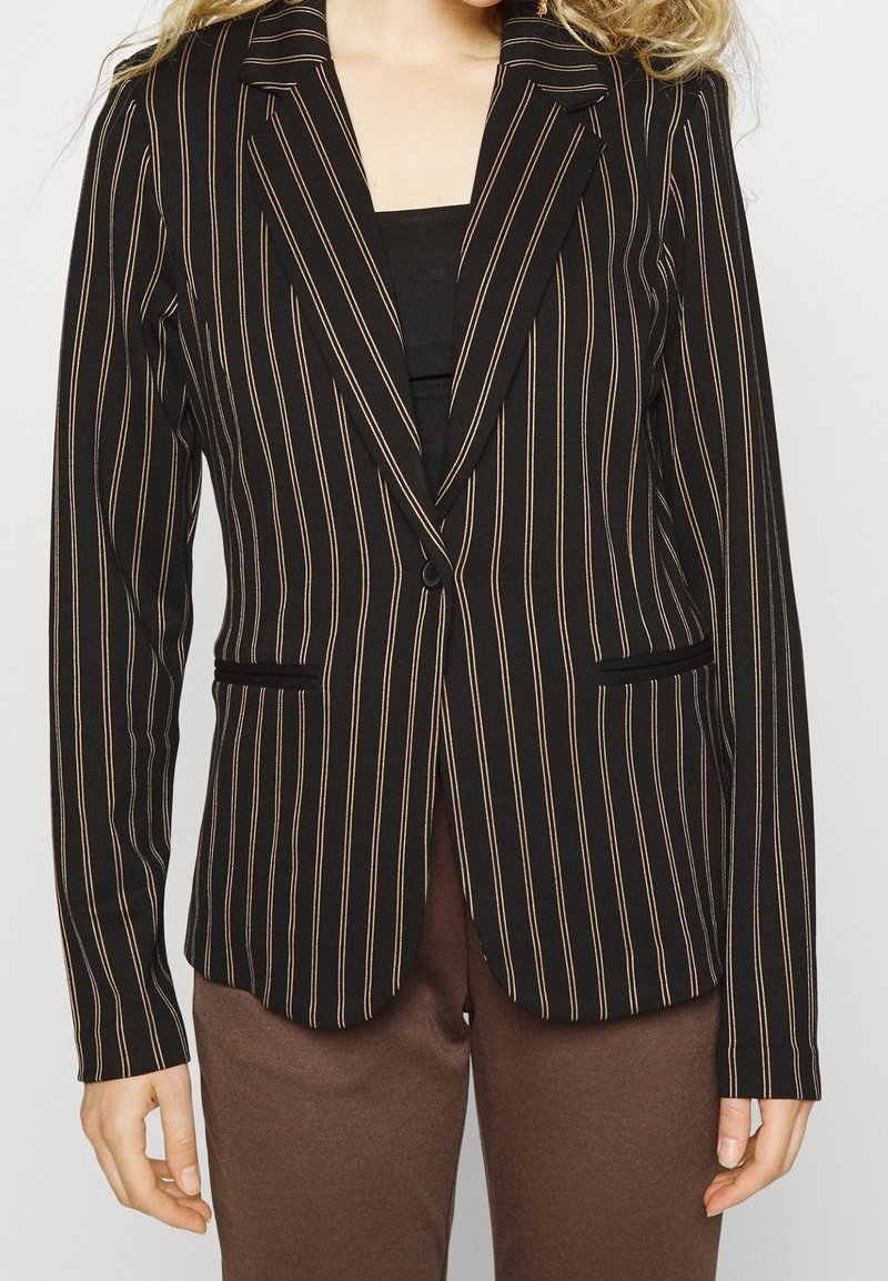 Veste noire à fines rayures verticales blanches, fermeture à un bouton, portée sur un haut noir, associée à un pantalon marron.