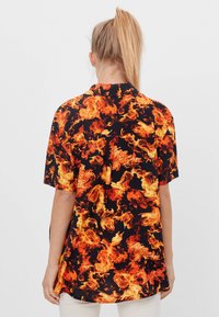 Kortärmad skjorta med en svart tygbas och ett mönster av lågor i nyanser av orange och gult, med en lös passform.