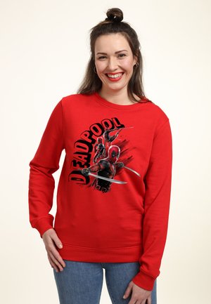 Uśmiechnięta kobieta z ciemnymi włosami związanymi w kok nosi czerwoną bluzę Deadpool z nadrukiem i niebieskie dżinsy na jednolitym jasnym tle.