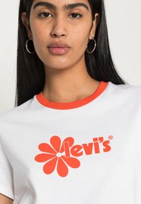 Bílý bavlněný tričko s červeným květinovým vzorem a logem "levi's" uprostřed, s kulatým červeným límečkem a krátkými rukávy.