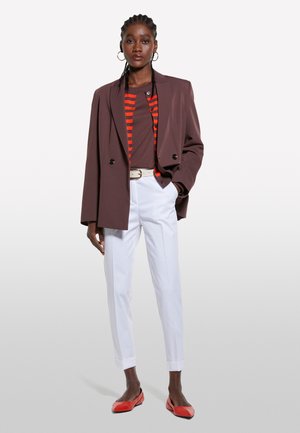 Blazer marrone doppiopetto sopra una camicia a righe rossa, pantaloni bianchi e scarpe rosse. Include orecchini a cerchio e una cintura chiara.