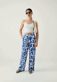 Λευκό τοπ με δαντέλα και κοντά μανίκια συνδυασμένο με παντελόνια φαρδιάς εφαρμογής σε μπλε και λευκό σχέδιο με φύλλα; μοντέλο που φοράει γυαλιά ηλίου και κρατά μια μικρή τσάντα.