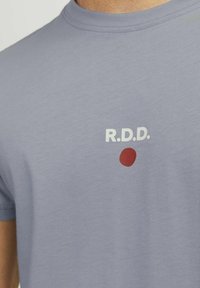 Tricou din bumbac de culoare violet deschis, cu text alb "R.D.D." și un cerc roșu pe față; guler rotund și mâneci scurte.