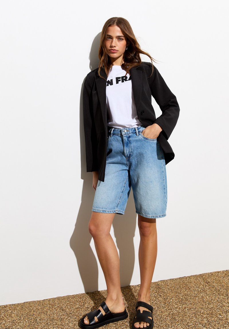 Blazer noir sur un t-shirt blanc avec du texte "FR", associé à un short en denim bleu clair et des sandales noires. Textures lisses, coupe décontractée.