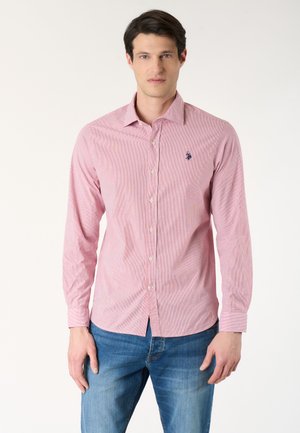Uomo che indossa una camicia rosa aderente a maniche lunghe con piccolo logo sul petto e jeans blu, in piedi davanti a uno sfondo semplice.
