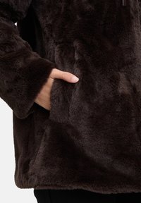 Giacca in pelliccia sintetica marrone con una texture morbida, dotata di tasca laterale e maniche ampie. Il materiale sembra essere soffice e spesso.