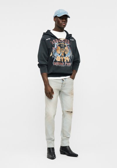 Jeune homme portant un sweat à capuche noir à motif, un jean clair déchiré, des bottes noires et une casquette de baseball bleu clair, debout avec une main dans la poche.