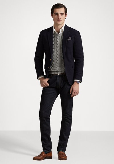 Blazer azul marino sobre un suéter de punto en gris, camisa blanca, jeans de mezclilla oscuros y mocasines marrones, con un pañuelo de bolsillo estampado.