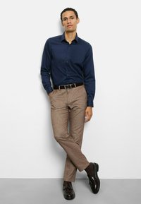 Marineblaue Langarmbluse mit Knopfleiste am Kragen, kombiniert mit hellbraunen, strukturierten Hosen und dunkelbraunen Loafers. Schlichtes, figurbetontes Design.