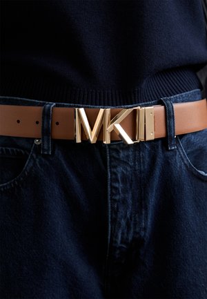 Brown leather belt featuring a gold-tone metal buckle with an 'MK' logo. Worn over dark denim. Subtle texture on the belt with visible holes.