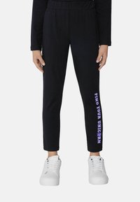 Zwarte sweatpants met een taps toelopende pasvorm en een elastische tailleband, met paars tekst "VIND JOUW EENHOORN" op het linkerpantalon. Gecombineerd met witte sneakers.