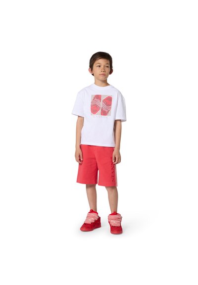 Garçon debout portant un t-shirt blanc à motifs, un short rouge avec des bandes latérales, et des baskets montantes rouges avec des lacets épais.