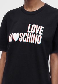 Μαύρο T-shirt με στιλιζαρισμένο κείμενο "LOVE MOSCHINO" με ένα λευκό σύμβολο καρδιάς στη θέση του γράμματος Ο στο στήθος.