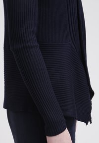 Gros plan d'une personne portant un cardigan côtelé bleu marine foncé à manches longues avec des motifs tricotés horizontaux et verticaux.