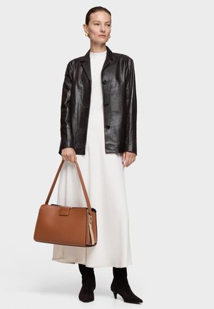 Donna che indossa una giacca di pelle nera, un lungo vestito bianco, stivali neri, che tiene una grande borsa marrone con doppi manici su uno sfondo neutro.