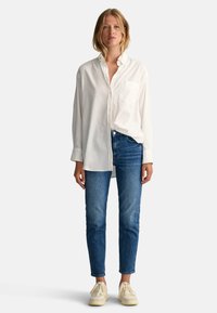 Wit overhemd met knopen en een borstzak, gecombineerd met blauwe denim jeans en crèmekleurige sneakers, staand tegen een effen achtergrond.