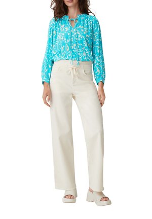 Femme portant un chemisier à manches longues turquoise à fleurs, un pantalon large crème taille haute et des sandales blanches à talon compensé et bout ouvert.