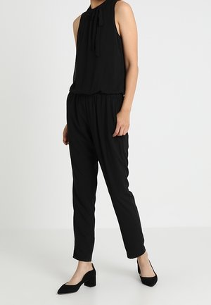 Femme portant un top sans manches noir avec un nœud au cou, un pantalon tapered taille haute noir et des chaussures à talons carrés noires, debout.